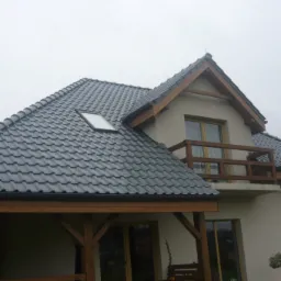 Dom z ciemnoszarą dachówką ceramiczną, drewnianą więźbą i oknem dachowym, sfotografowany w pochmurny dzień.